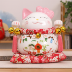 Maneki-Neko de cerámica blanco con lazo rosa, campanas doradas y cojín rojo floral