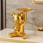 Figura decorativa metálica en forma de oso dorado que sostiene una bandeja, colocada sobre un mueble junto a una pared gris