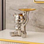 Figura decorativa metálica en forma de oso plateado que sostiene una bandeja dorada, colocada sobre un mueble junto a una pared gris